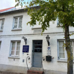 Weißes Backsteingebäude mit grauer Eingangstür, historischen Laternen und Straßenbaum
