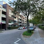 Wohnstraße mit modernen Mehrfamilienhäusern, Balkonen, Gehweg und Straßenbäumen
